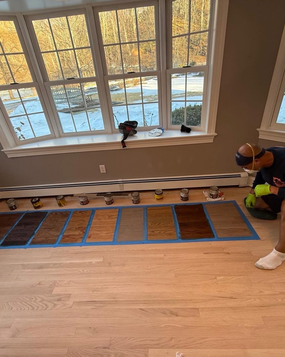 Stain color samples taped off on a bare oak floor beside a bay window