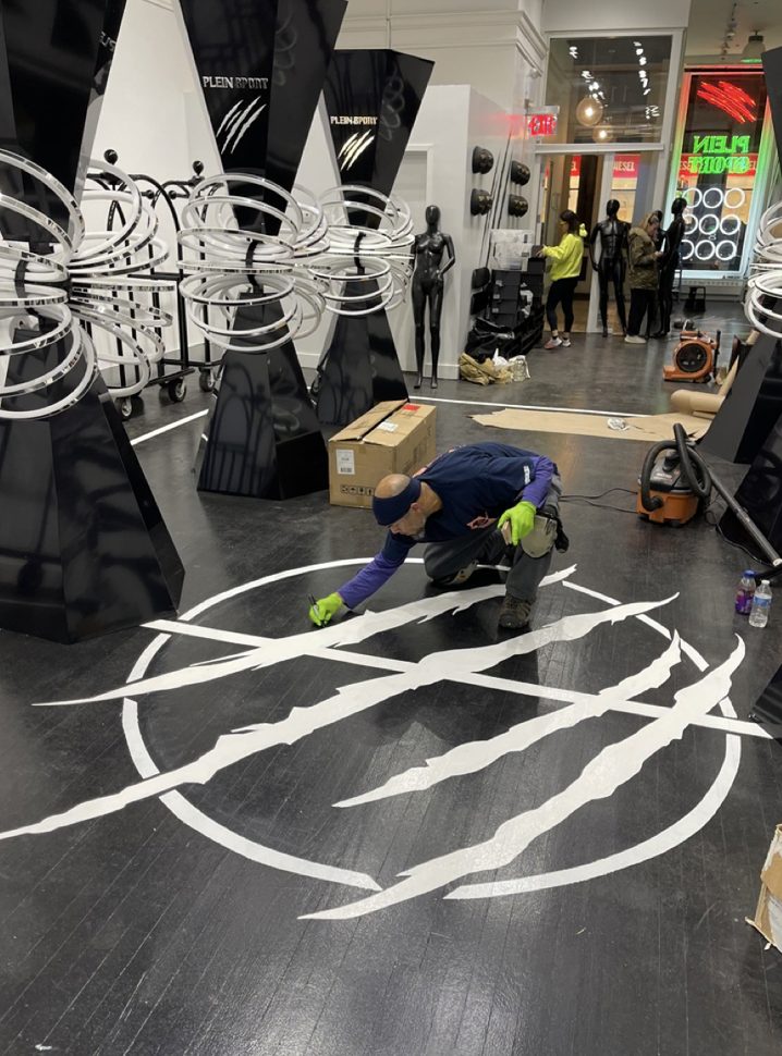 Kwazi kneeling to paint a white claw-mark graphic onto a black wood floor in a retail store