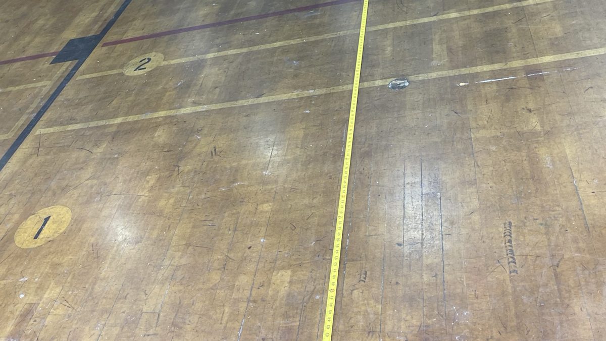 Worn, scuffed gym floor before refinishing, with a tape measure laid across it