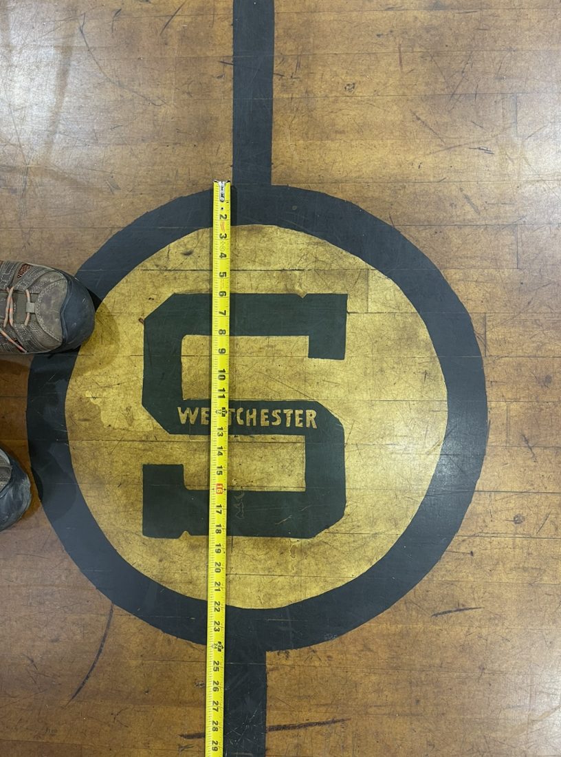 Old worn S Westchester logo painted at center court, with a tape measure laid across it
