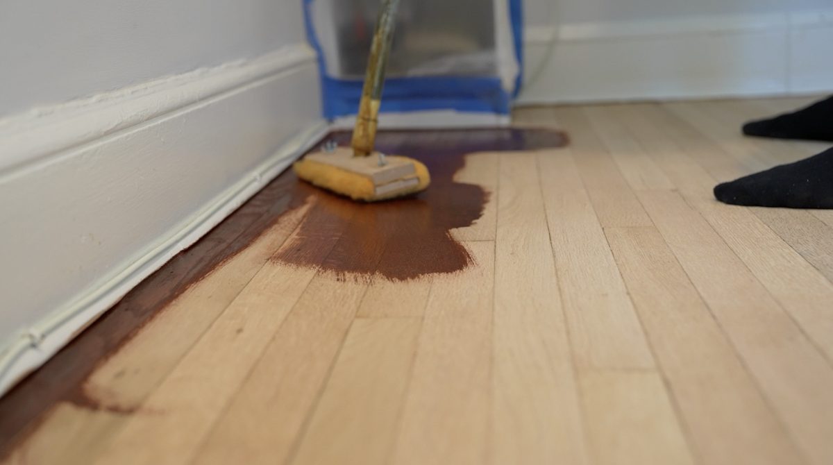 Applying stain to bare oak flooring with a lambswool applicator beside the baseboard